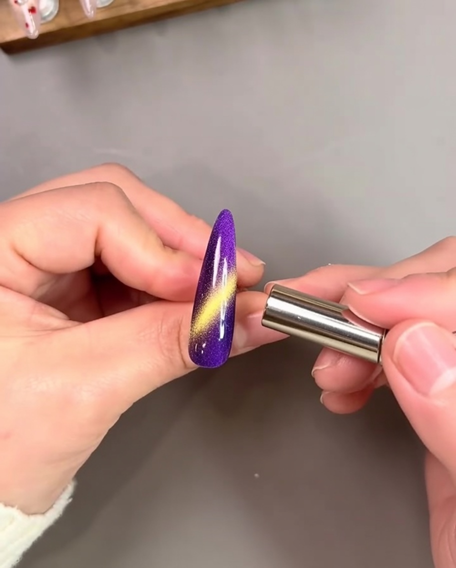 A hand holding a cylindrical magnet close to a vibrant purple painted nail, activating a bright gold cat-eye streak.