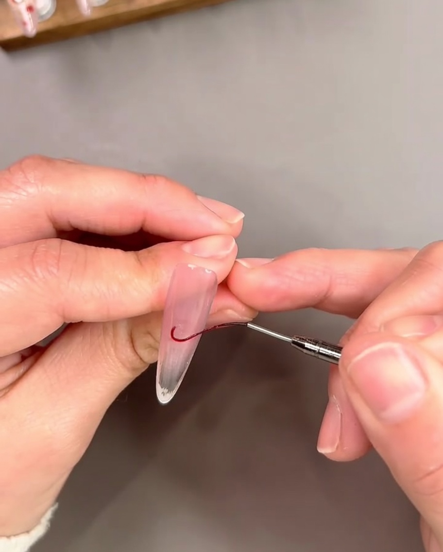 A fine liner brush drawing a thin curved red gel line near the free edge of a nail to outline a French tip.