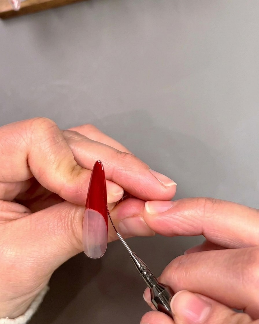 A thin liner brush sharpening the inner edge of a deep red V-shaped French tip on a nail.