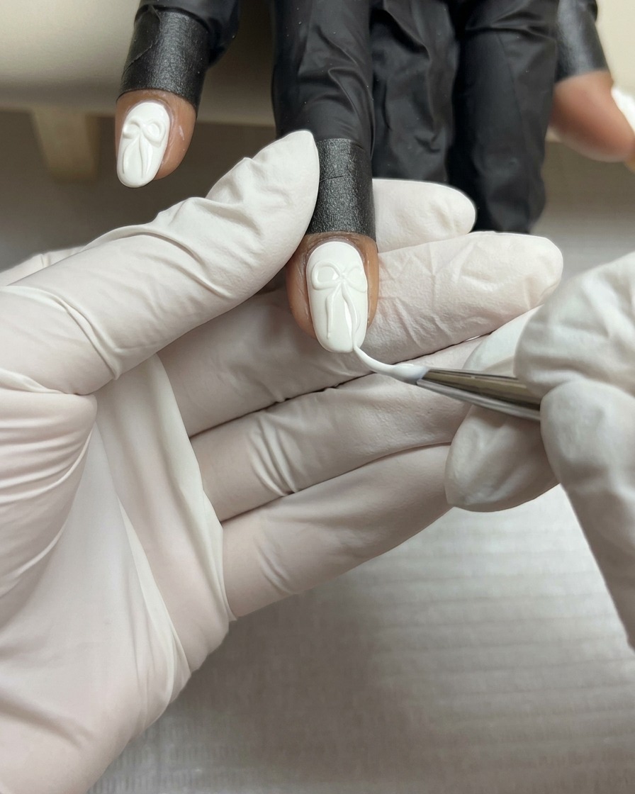 A close-up of a fine liner brush adding thin white lines to the surface of a 3D gel bow on a white nail.