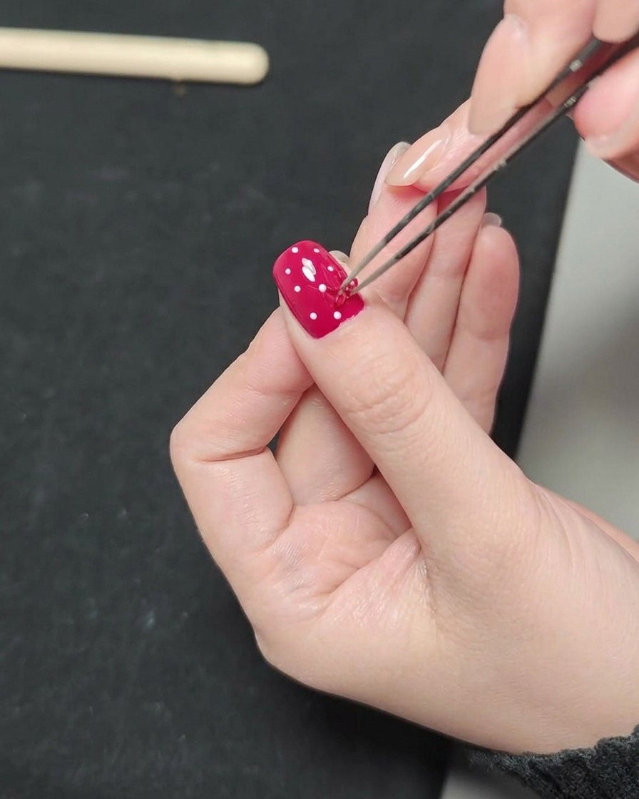 A close-up view of precision tweezers holding a small red 3D bow charm over a painted nail surface.