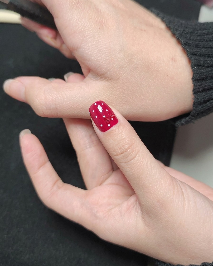 A nail polish brush applying a glossy clear top coat over a red nail with white polka dots and a red bow charm.