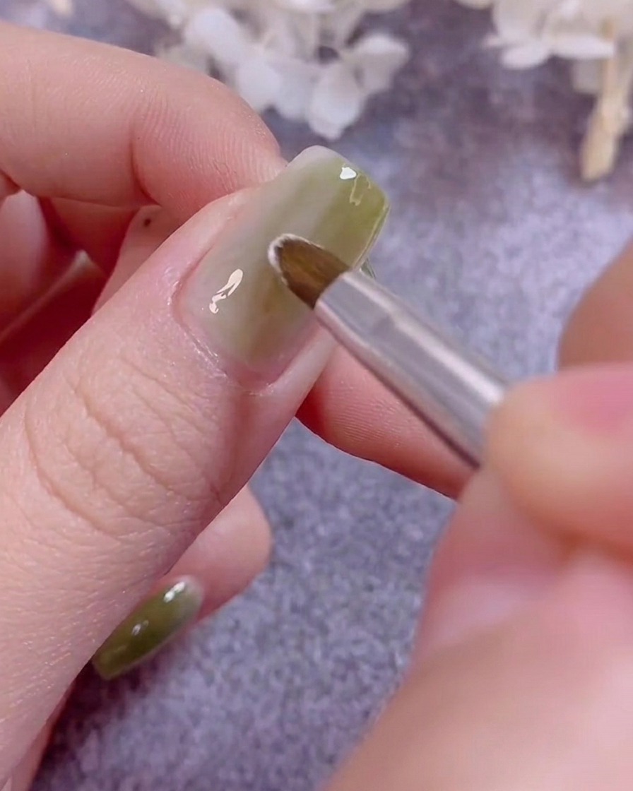 A thin nail art brush painting delicate white flower petals on a green gel-coated nail.