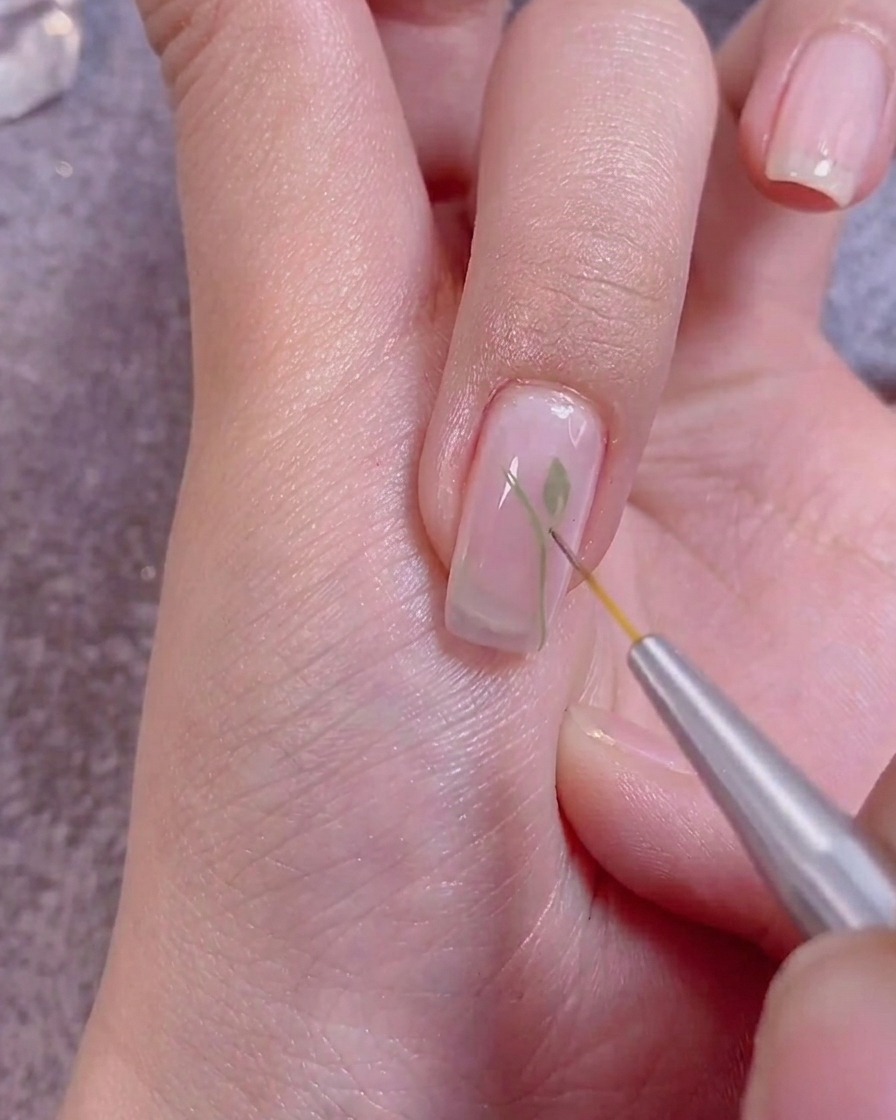 A close-up shot showing a fine brush adding white detail to green painted leaves on a nail.