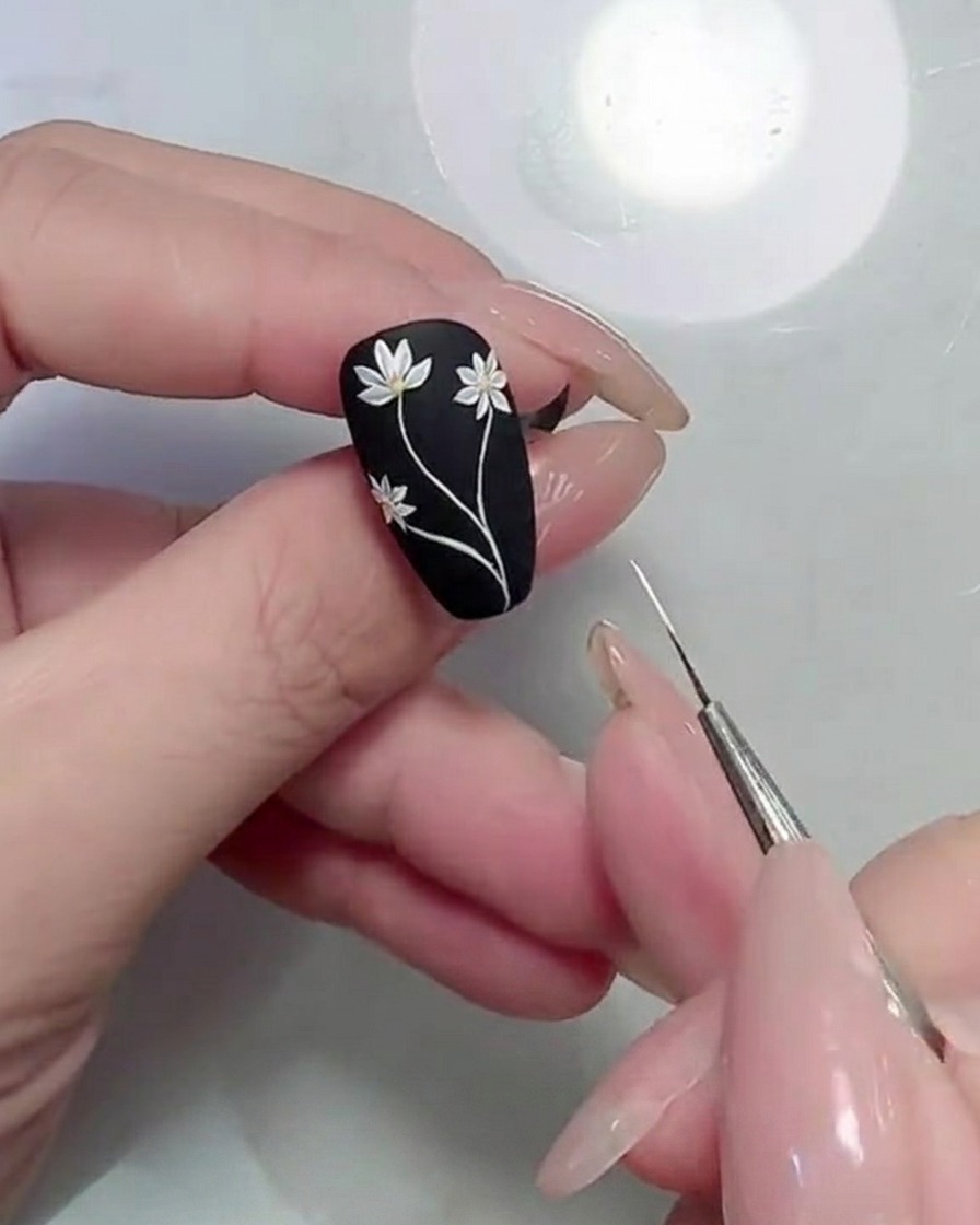 A close-up of a detail brush applying green leaf shapes next to white daisy stems on a matte black nail.