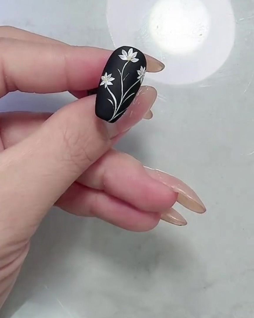 A macro shot showing the completed daisy and leaf design on a matte black nail, focusing on the refined petal and leaf edges.