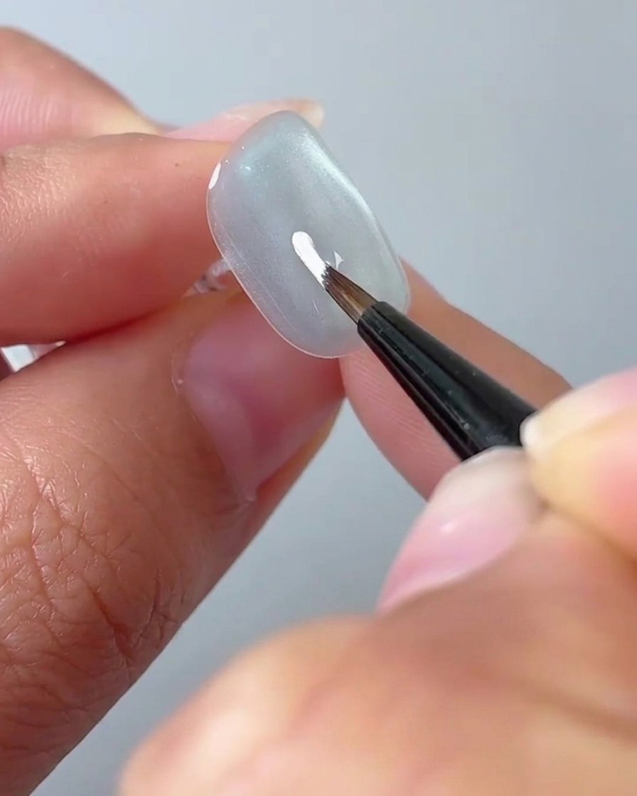 A fine liner brush drawing a white petal on a light grey nail surface.