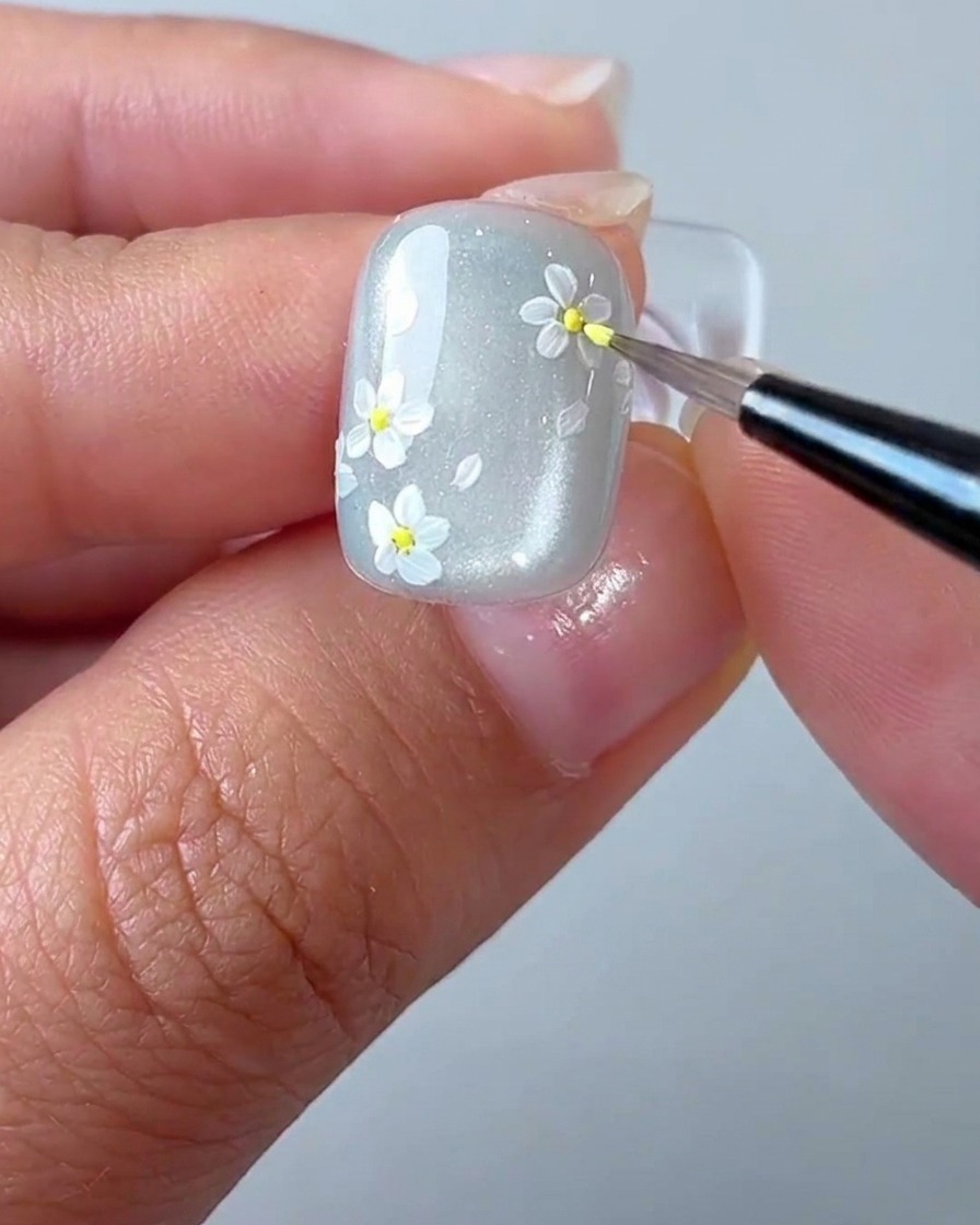 A detail brush applying a tiny yellow dot to the center of a white daisy on the nail.