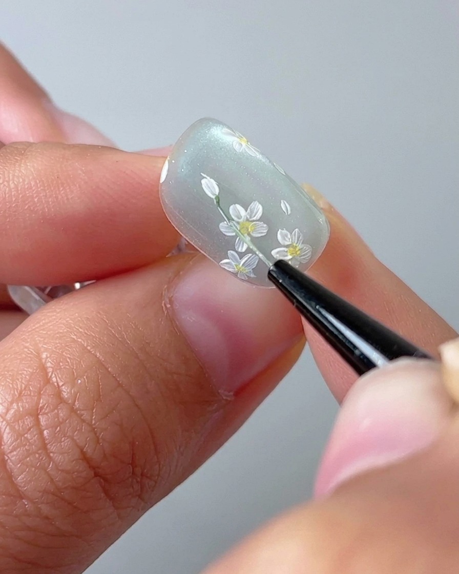 A close-up of a nail brush drawing a thin green stem on a cat-eye gel nail base.