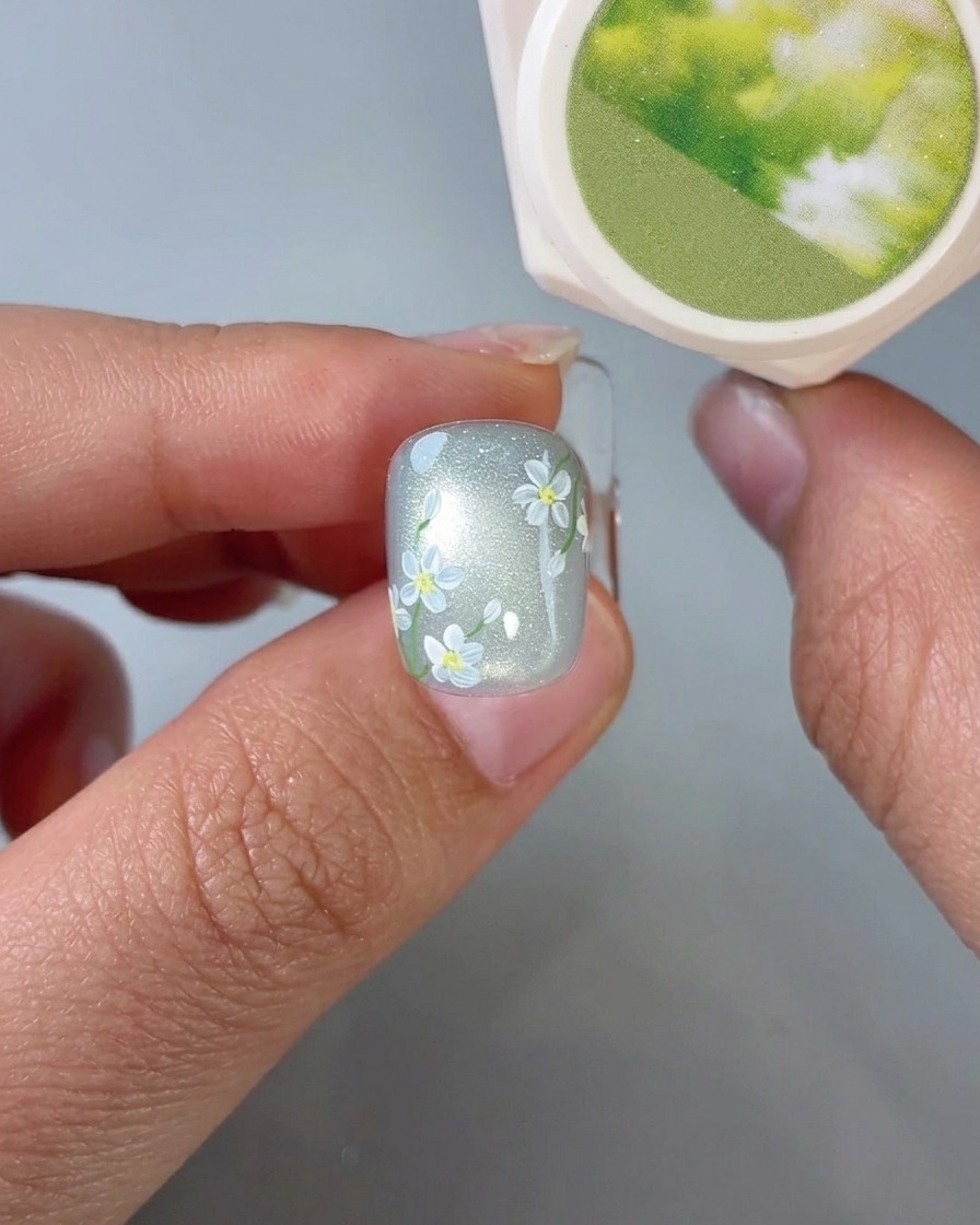 A close-up view of a brush applying small green leaf details onto a nail with daisy art.