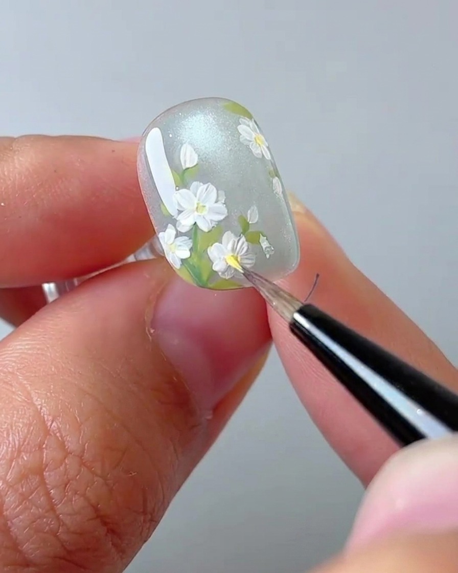 A close-up shot of a nail artist using a brush to place a yellow dot in the center of a white daisy.