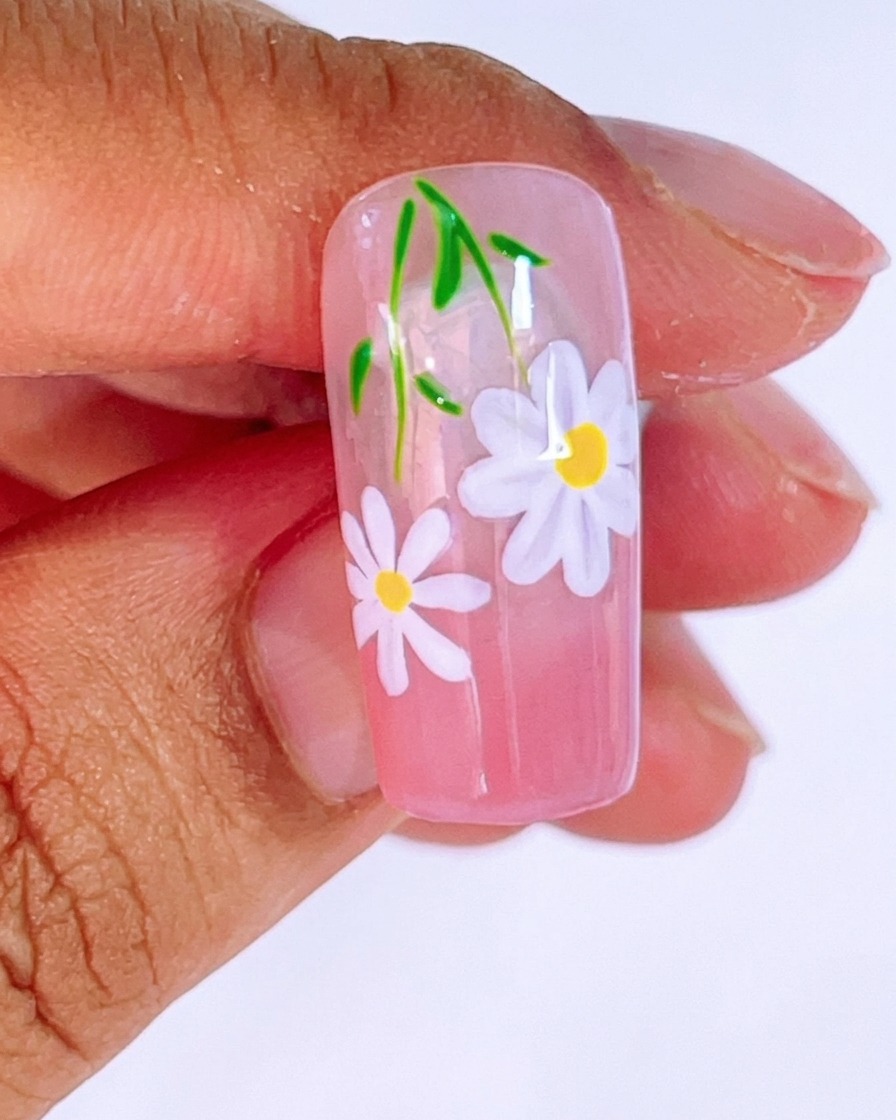 A close-up of a pink gradient nail being painted with a second, smaller daisy using white gel and a fine liner brush.