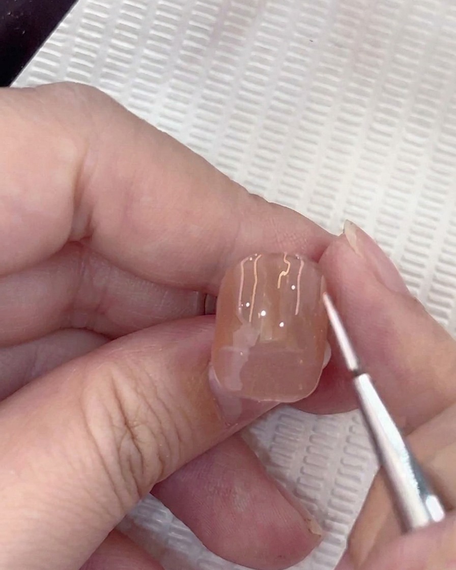A close-up of a liner brush applying a small white guide dot to the center of a nude gel nail.