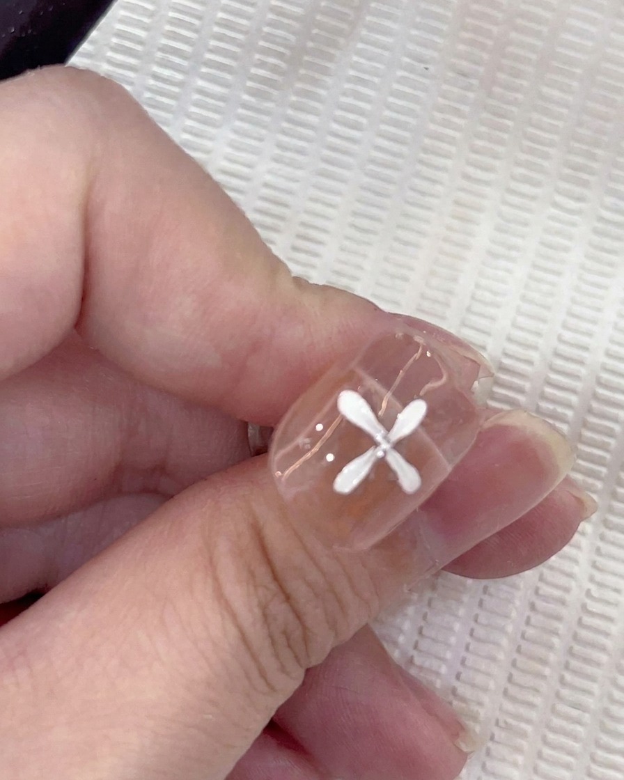 A close-up of a nail with five white petals painted in a circular pattern, with a brush adding a sixth petal.