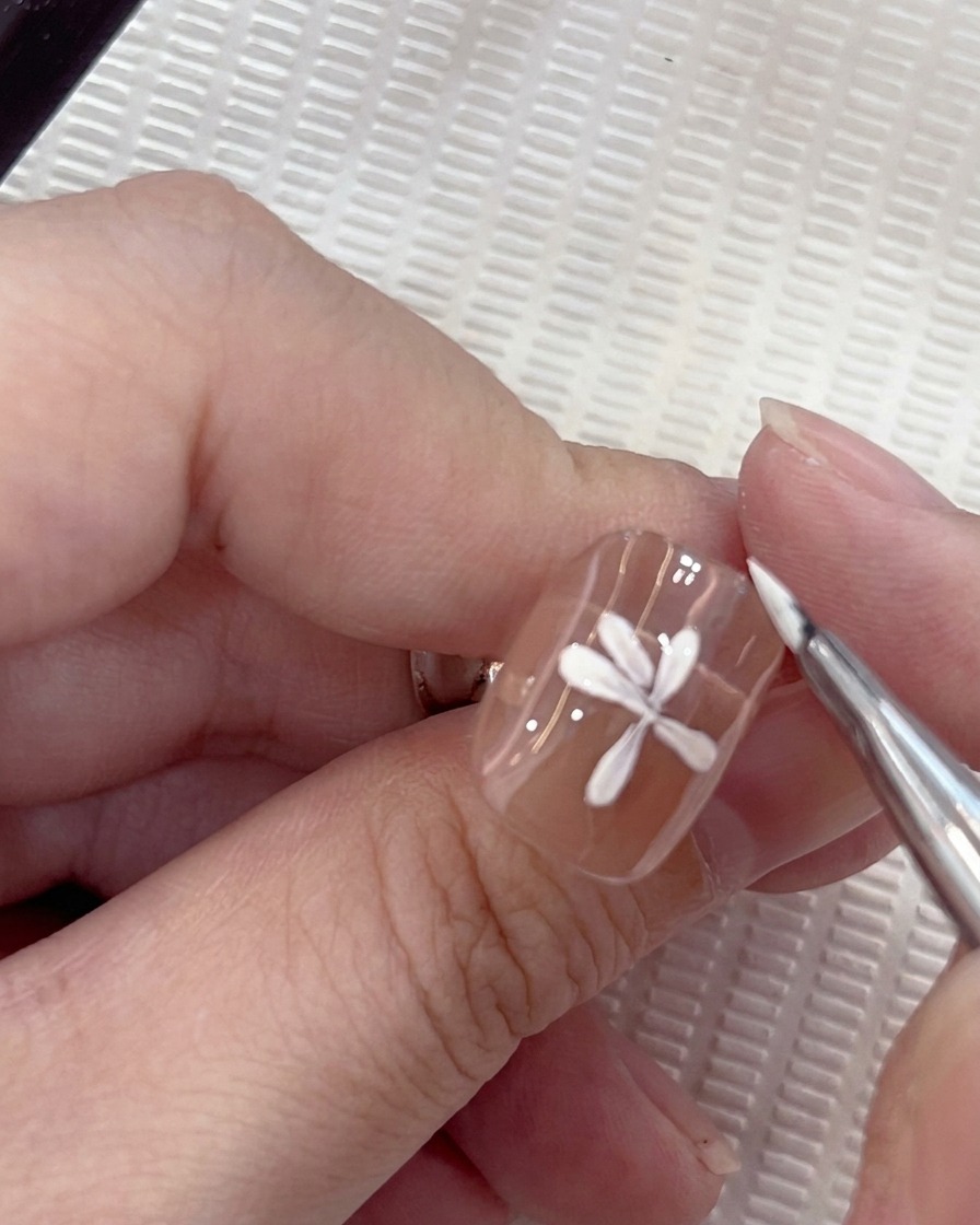 A close-up of a white daisy nail art design where a seventh petal is being carefully placed by a round brush.