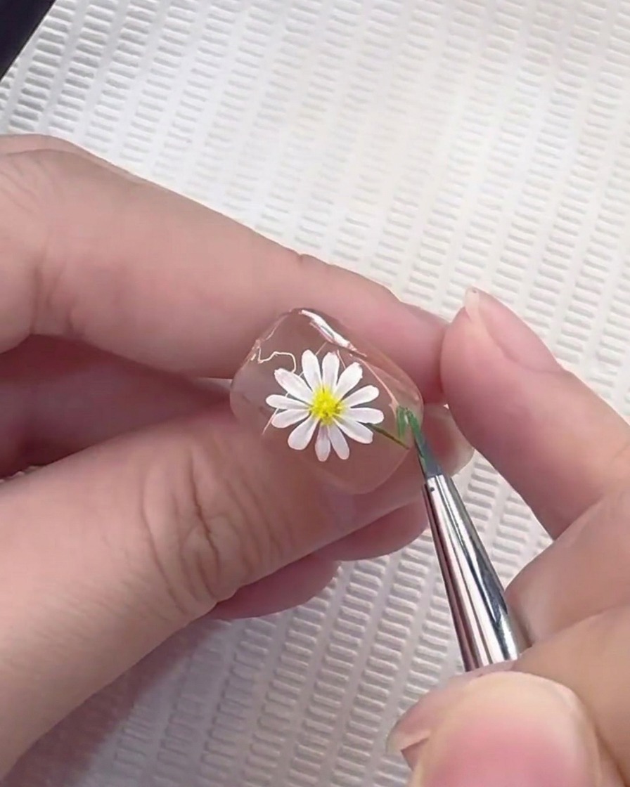 A liner brush pulling a thin green line to form a stem on a daisy-decorated nail.