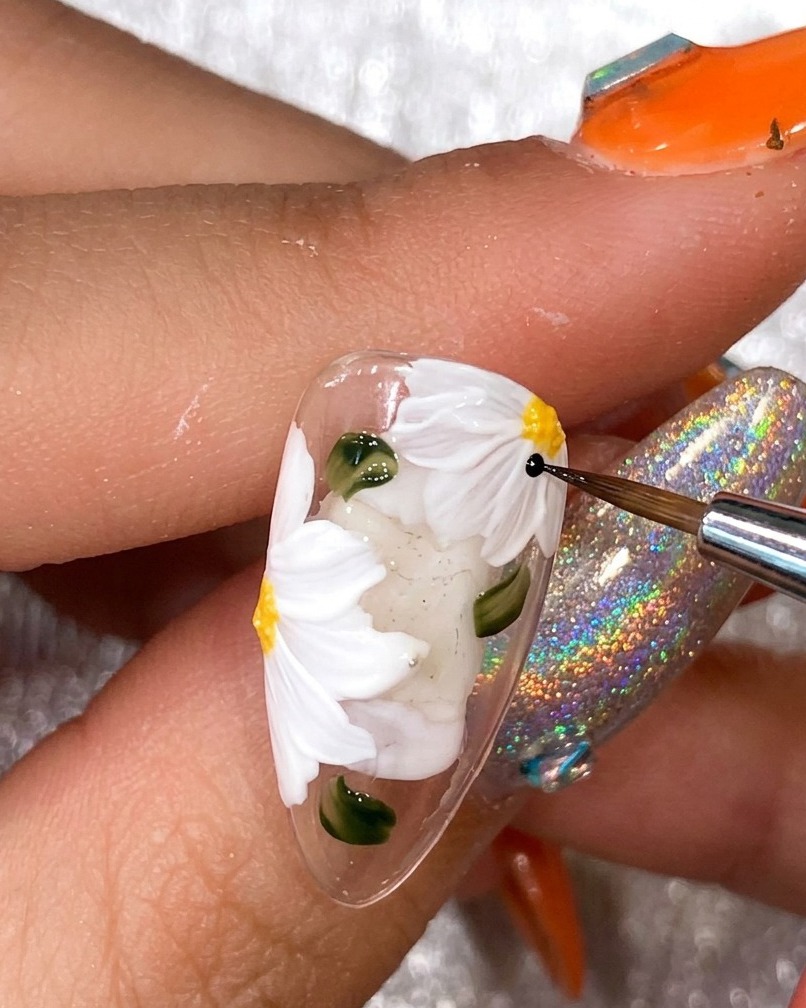 A close-up view of a nail artists hand using a thin black liner brush to outline white daisy petals on a nail tip.