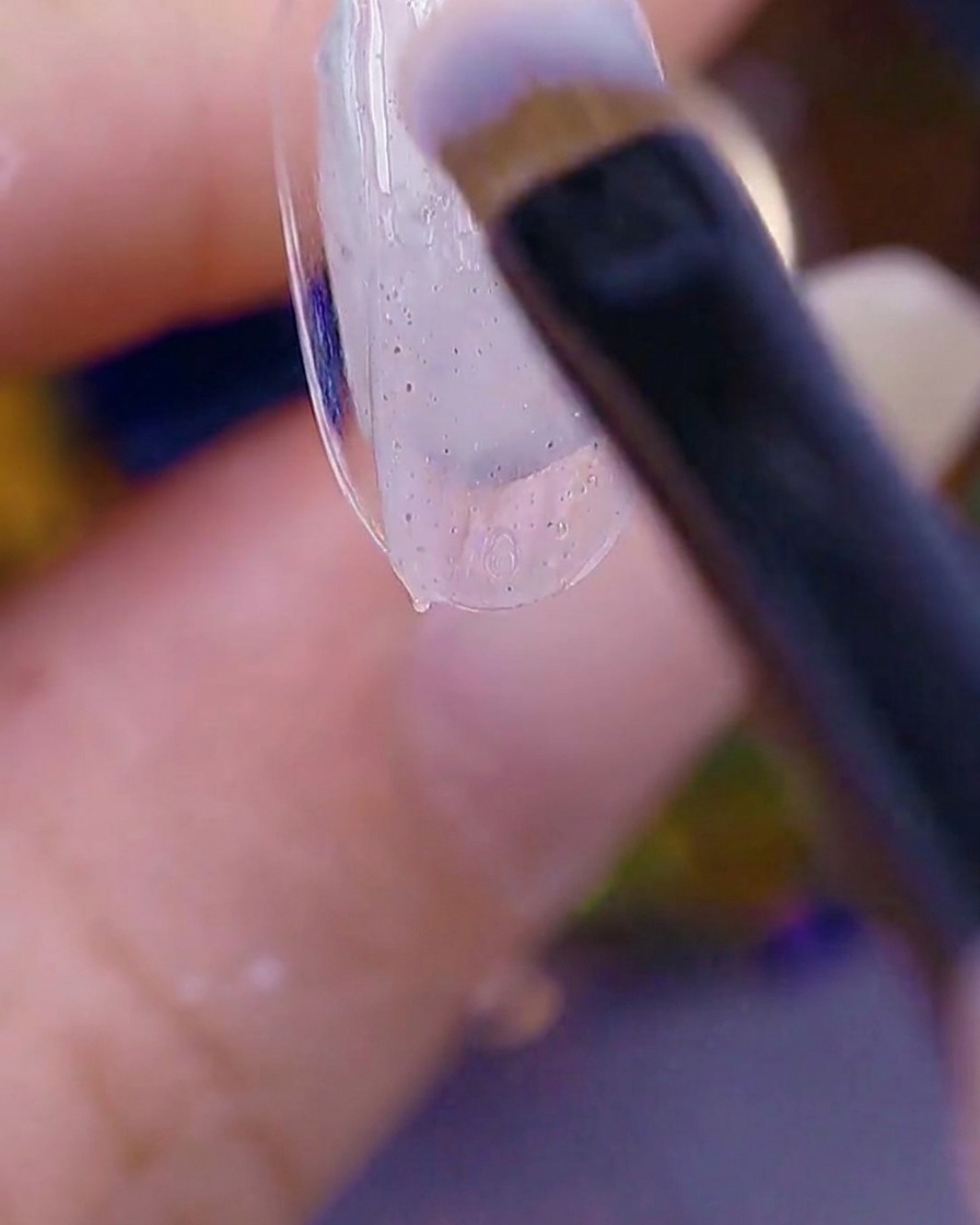 A close-up view of a brush applying a sheer, sugar-textured gel to the tip of a natural nail.
