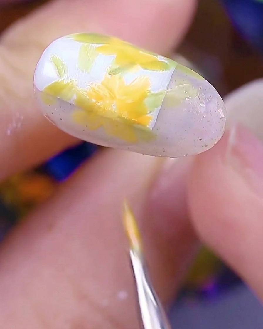 A close-up view of the nail showing multiple yellow-orange daisy flowers painted over the leaf bases, with the brush finishing the last petals.
