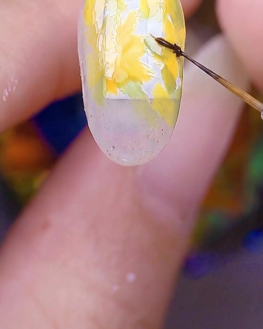 A close-up of a nail showing the yellow flower petals with a fine brush adding tiny brown dots to the very centers.