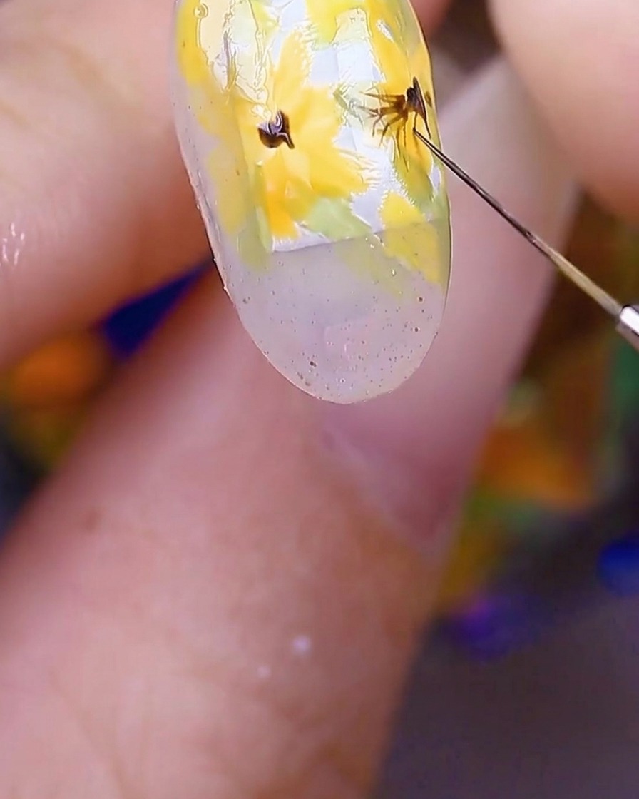 A close-up of the nail art showing a brush pulling fine, thin dark brown lines from the center of the yellow flowers outward into the petals.