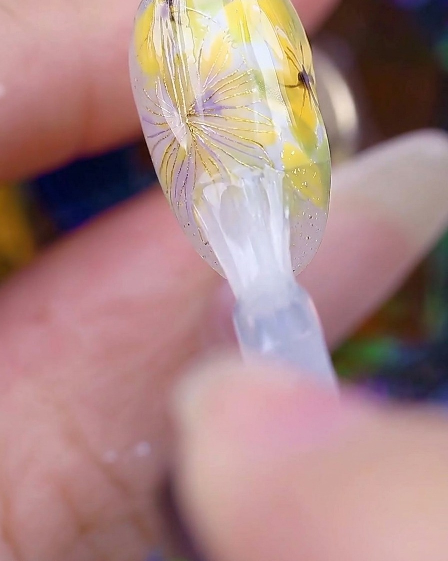A brush applying a clear, glossy top coat over the completed daisy nail art, making the surface look smooth and shiny.
