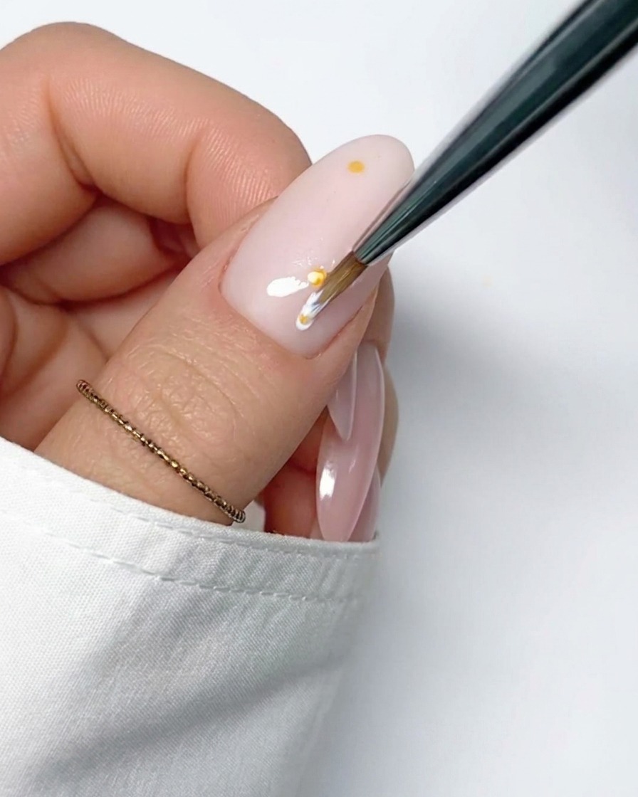 A detail brush applying a thin white stroke of gel starting from the yellow center dot, forming the first petal of a daisy.