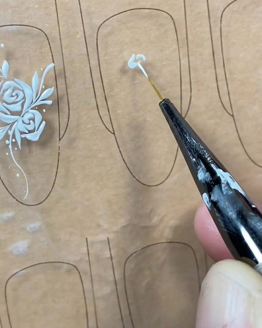 A close-up of a liner brush applying a small dot of white gel to the center of a clear nail tip.
