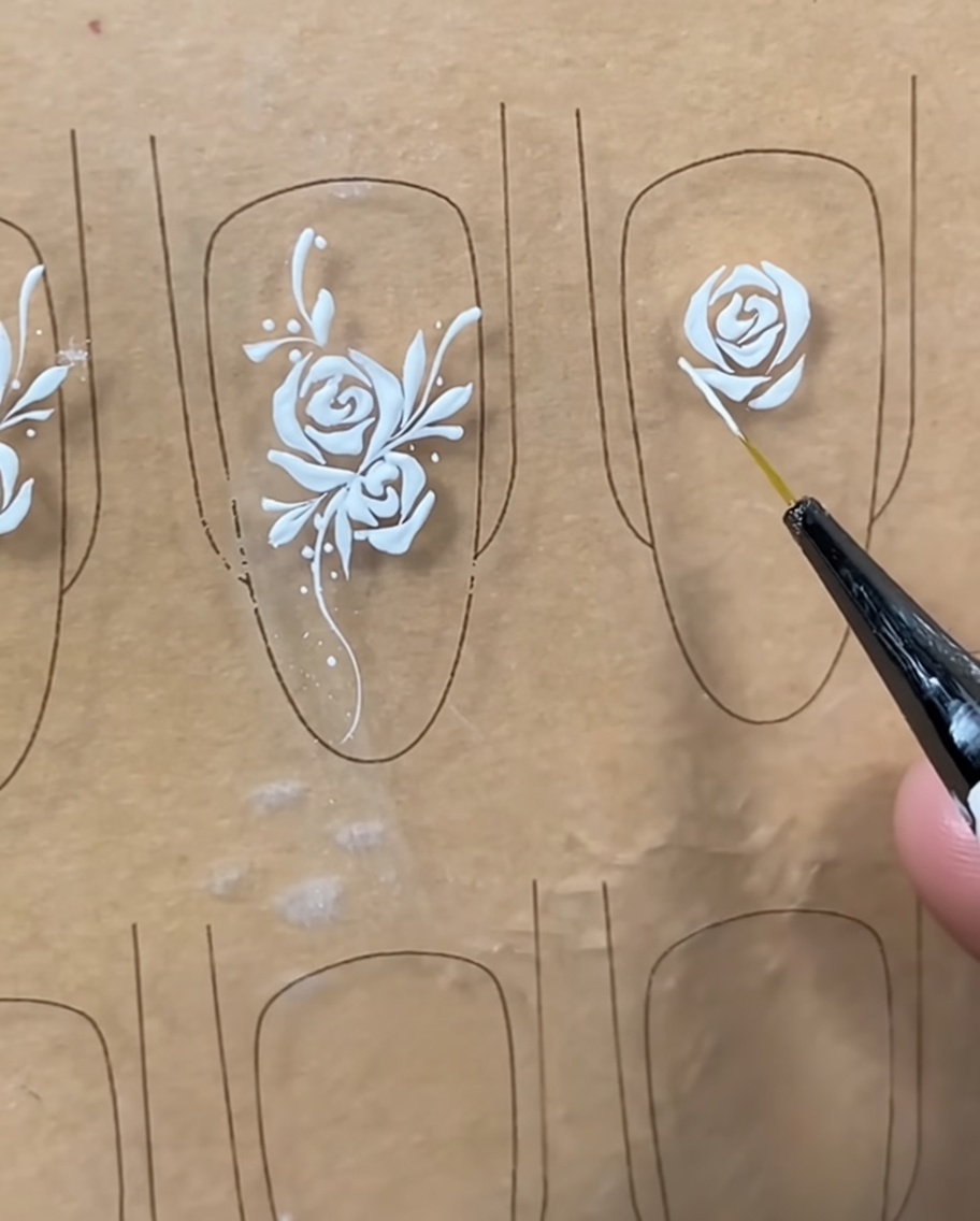 Close-up of a liner brush adding fine white details to the outer petals of a rose design on a nail tip.