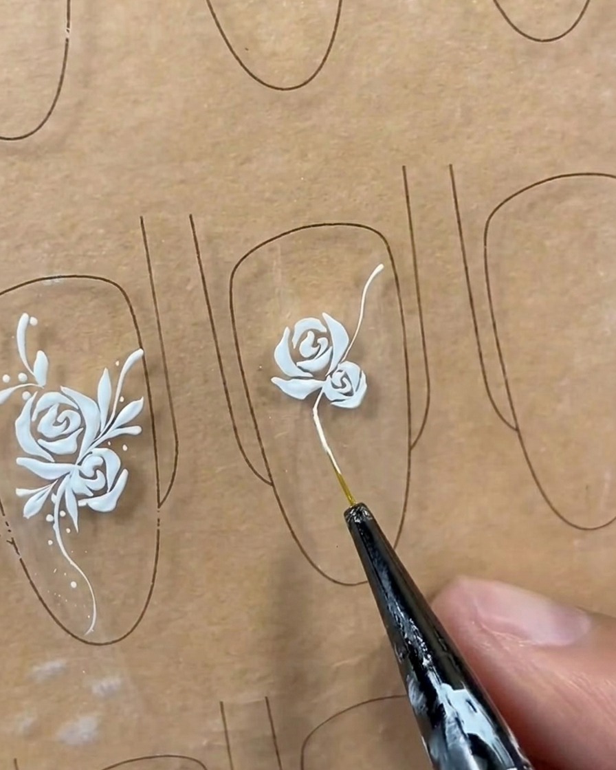 A long, sweeping white vine line being drawn from the base of a rose design on a nail tip using a liner brush.