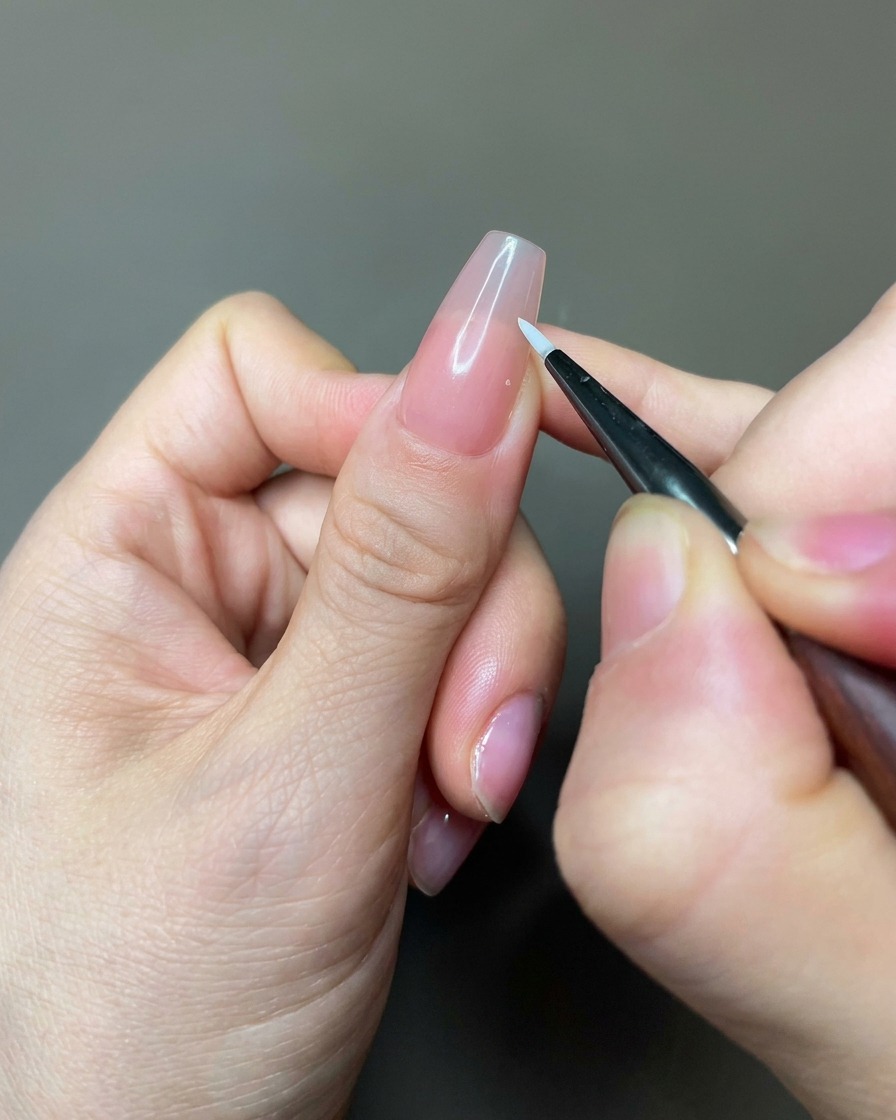 A close-up of a fine liner brush applying white gel to create petal shapes on a nude-colored nail.