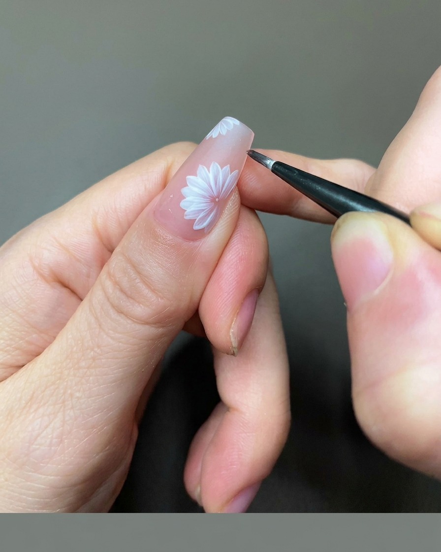 A close-up view of a nail technician using a fine liner brush to add golden gel details to the center of a white daisy on a nude nail.