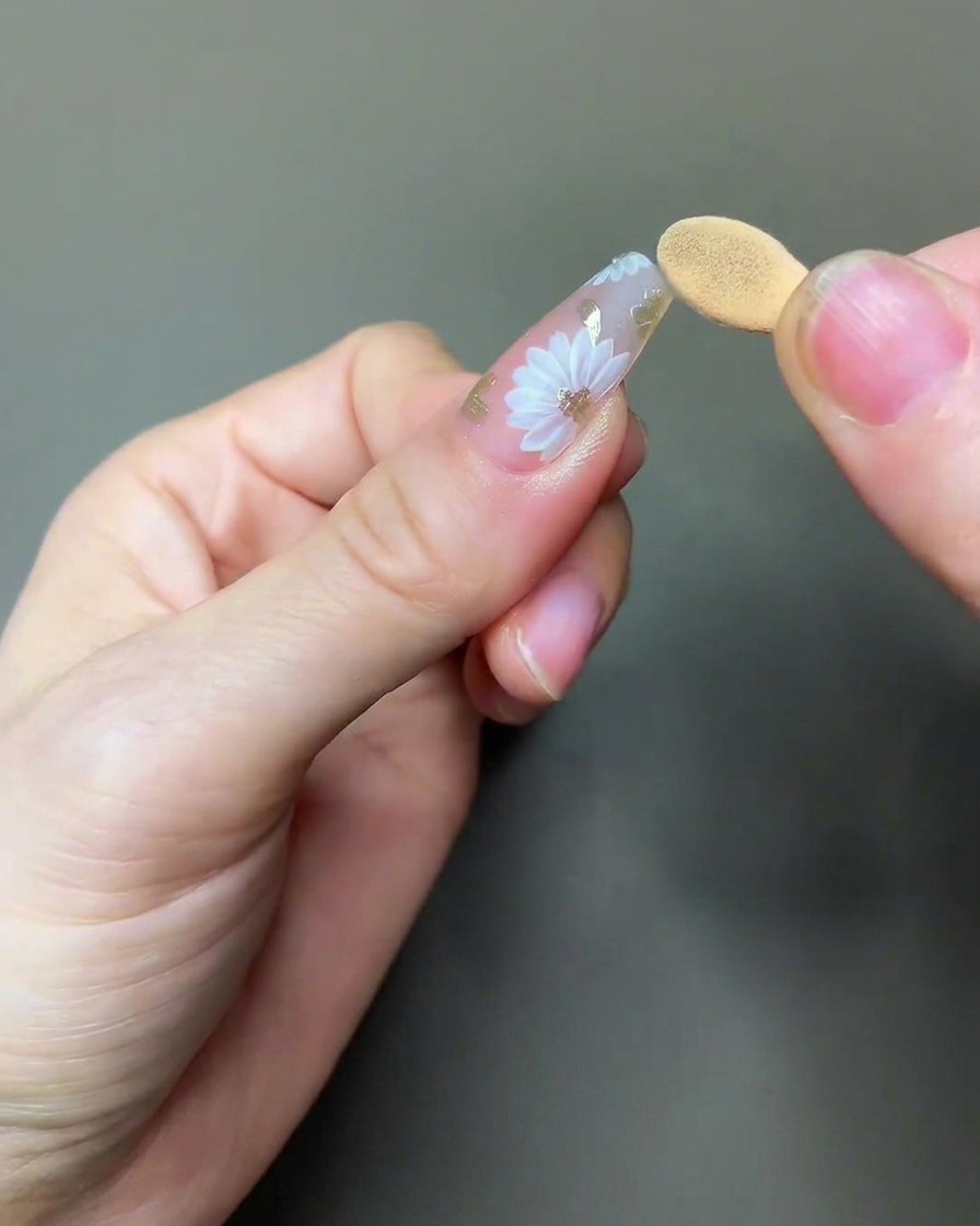 A brush applying a clear, glossy top coat over the completed daisy and chrome nail art.