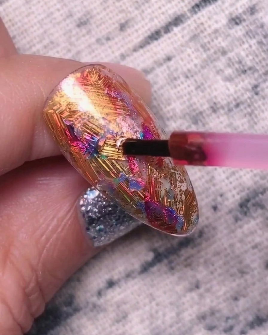 A small brush applying a pigmented red-purple gel to a textured nail surface covered in metallic foils.