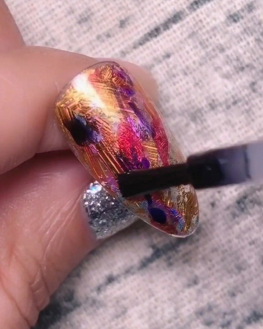 A close-up of a nail being dotted with blue-purple gel, showing the colors beginning to blend and expand into the base layer.