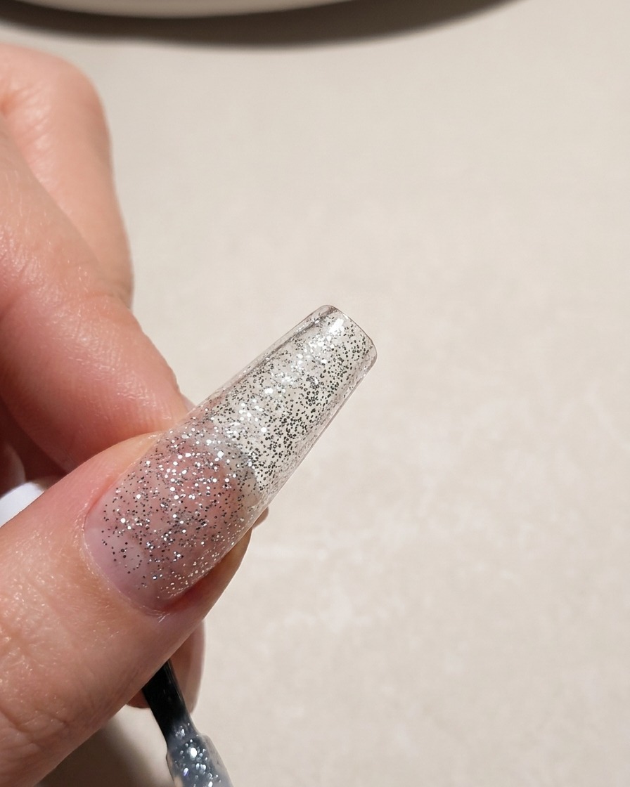 A close-up view of a nail tip being coated with a silver glitter gel polish.