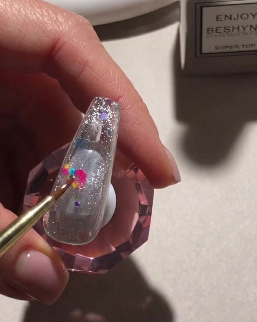A detail brush placing vibrant, multicolored round sequins onto a glittery nail surface.