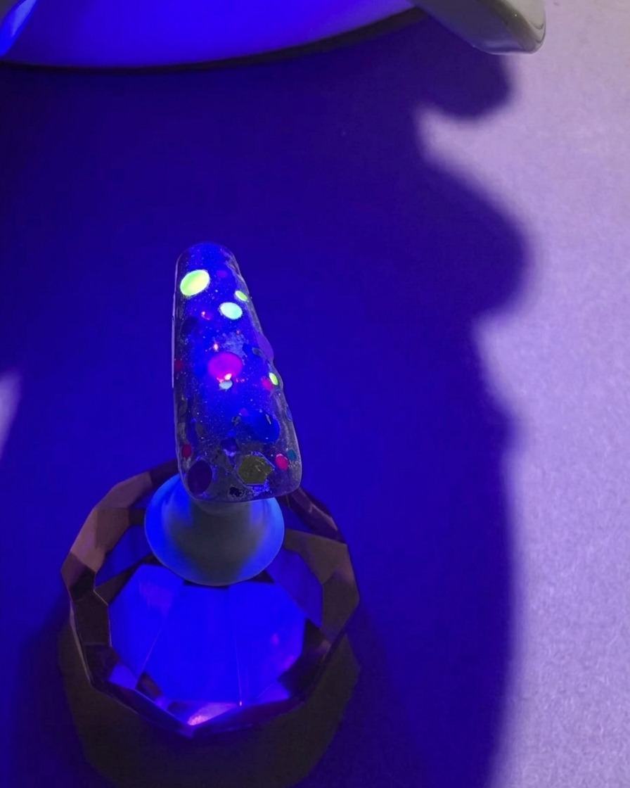 A nail with colorful sequins under the purple light of a UV lamp, mid-curing process.