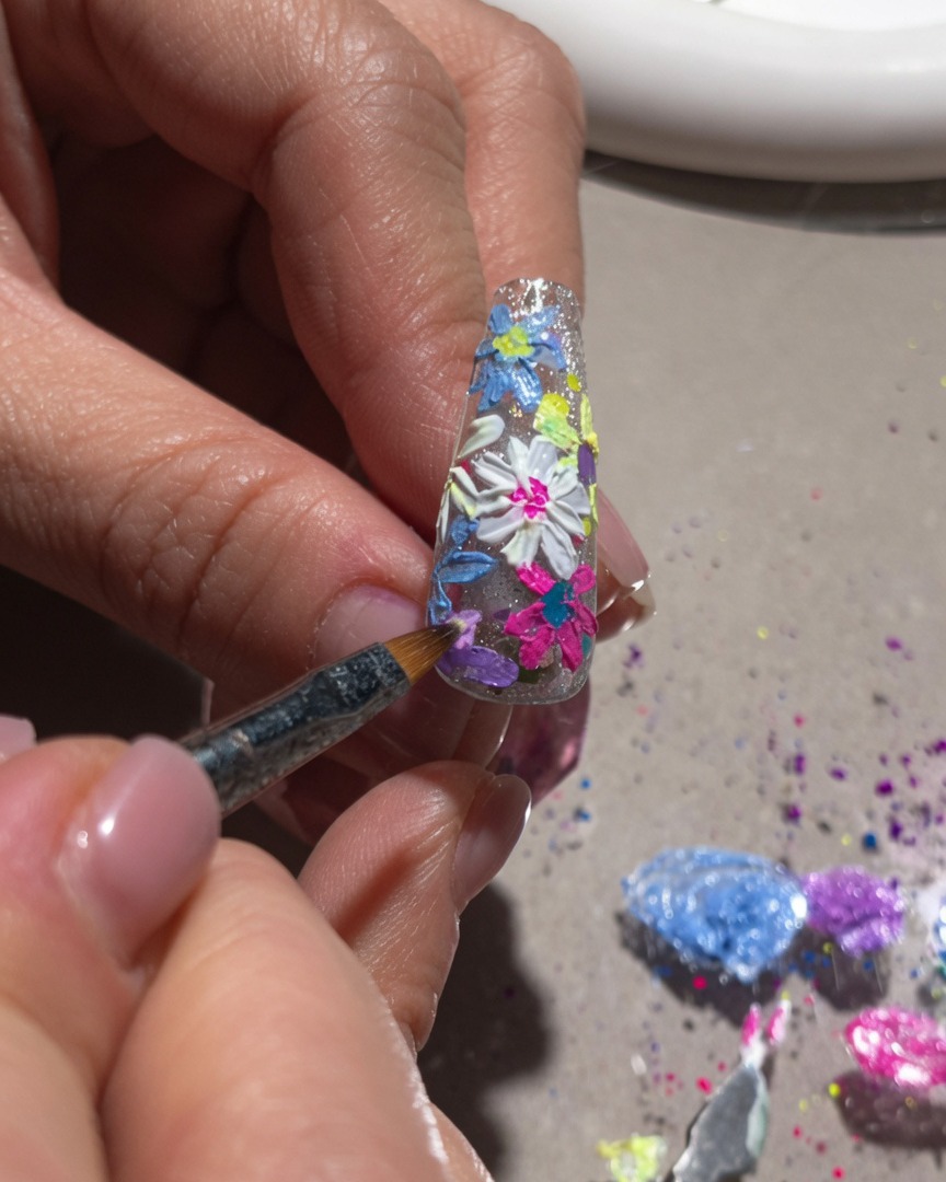A close-up view of a fine brush adding white highlights and center details to a 3D floral nail design.