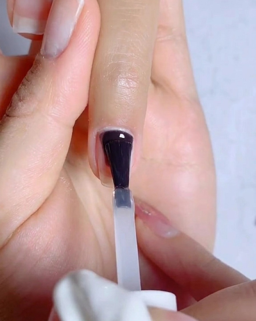 A close-up of a brush applying a clear, thin layer of gel base coat onto a natural, prepped fingernail.
