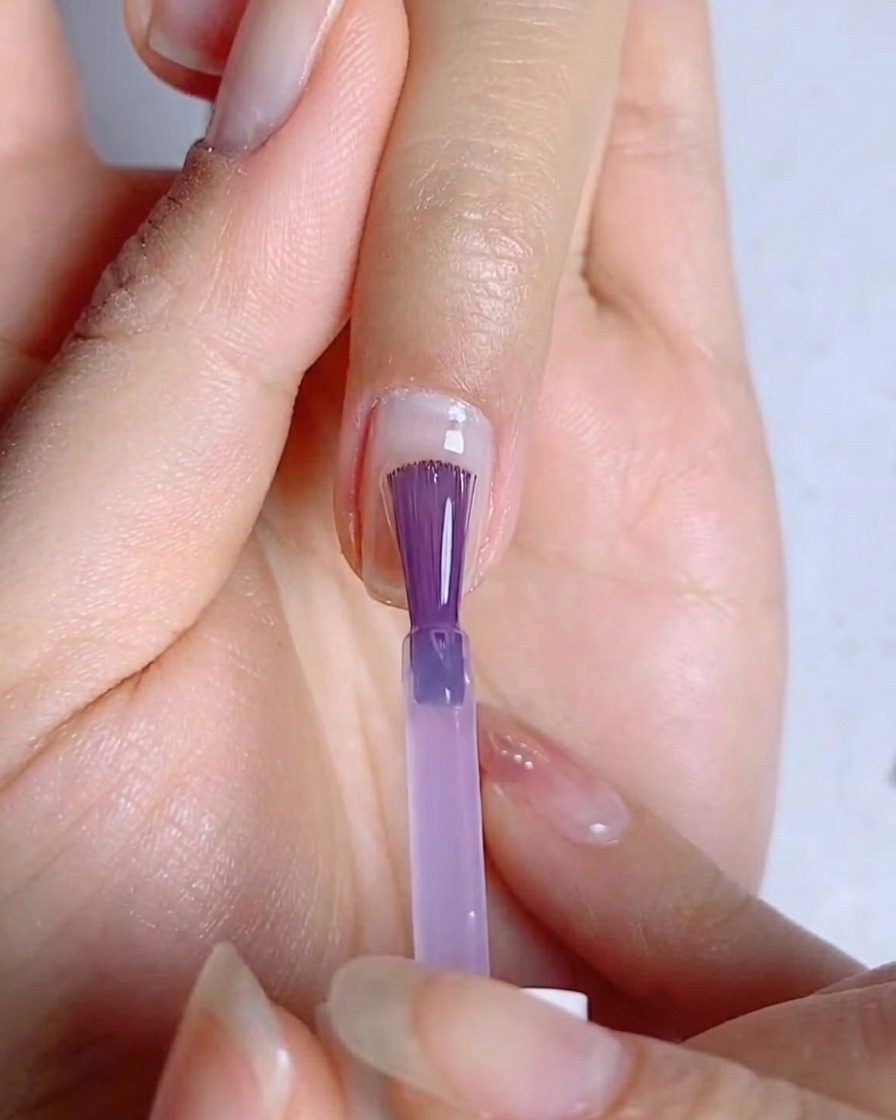 A nail brush spreading a thin, even layer of blue-purple gel polish over the nail surface.