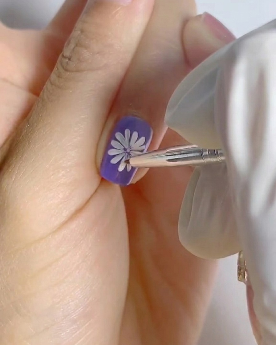 A finger with the completed white daisy art on a purple base placed under a small professional nail lamp for curing.
