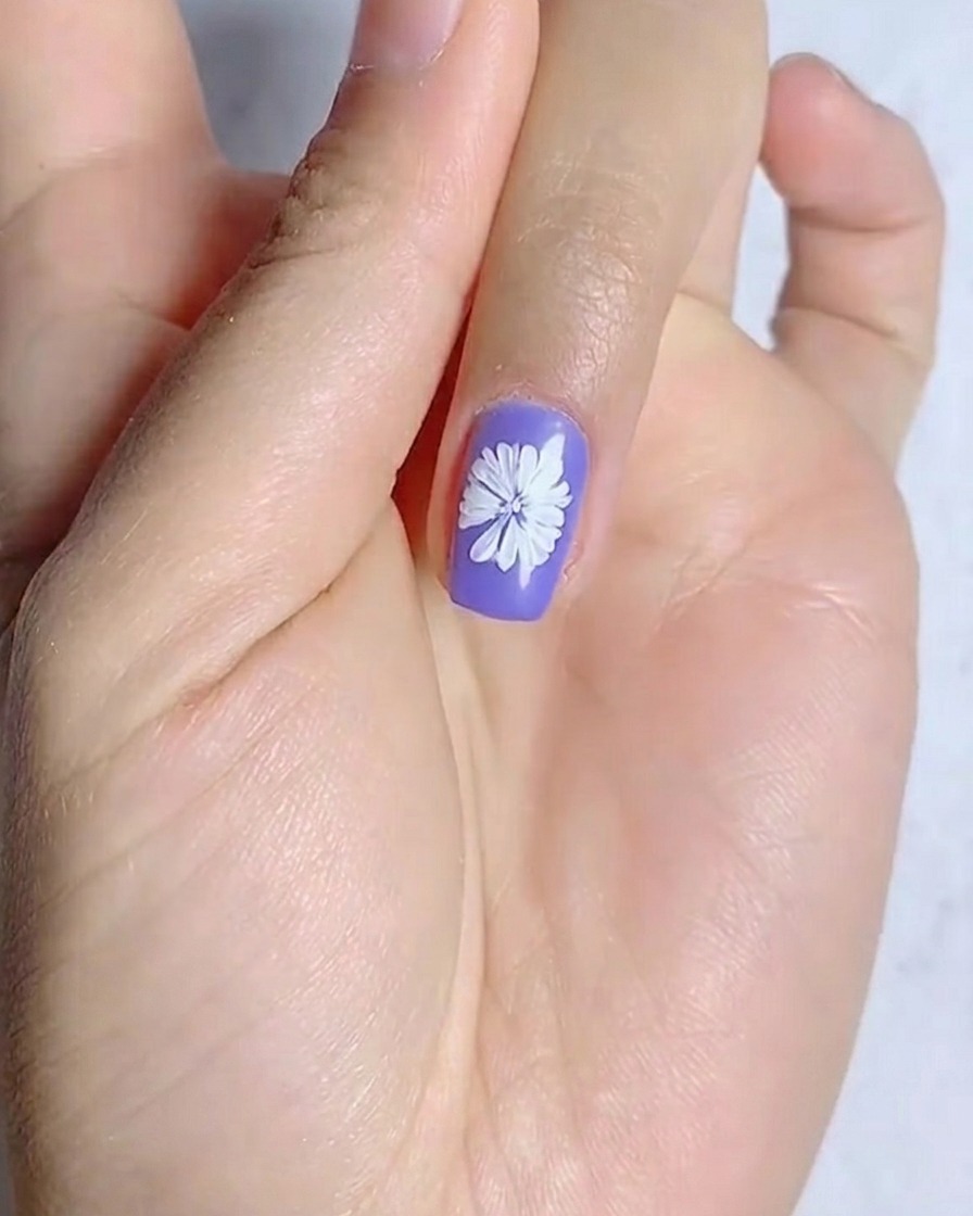 A close-up view showing a yellow dot being applied to the center of a white daisy painted on a purple nail.
