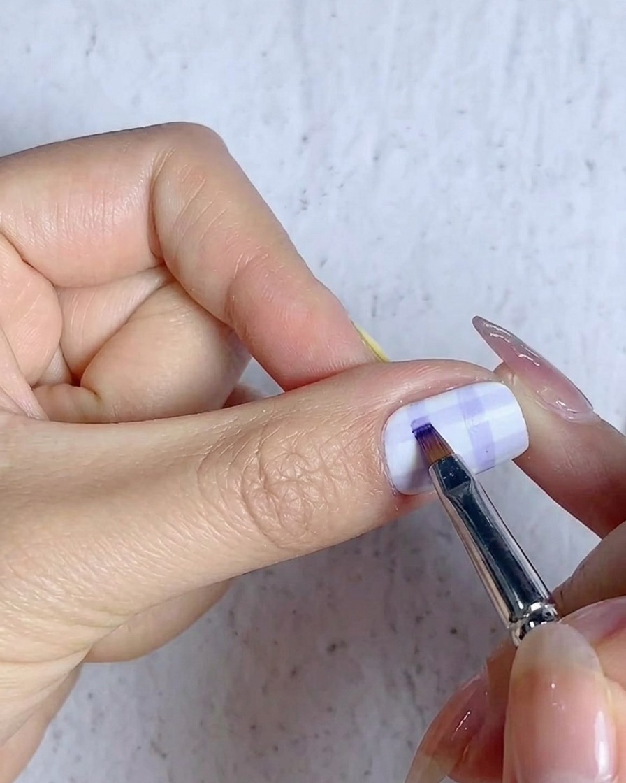 A fine brush filling in the grid intersections on a plaid nail design with blue gel.