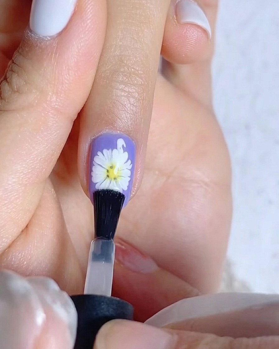 A brush applying a coat of matte top gel to a purple nail featuring a white daisy design.