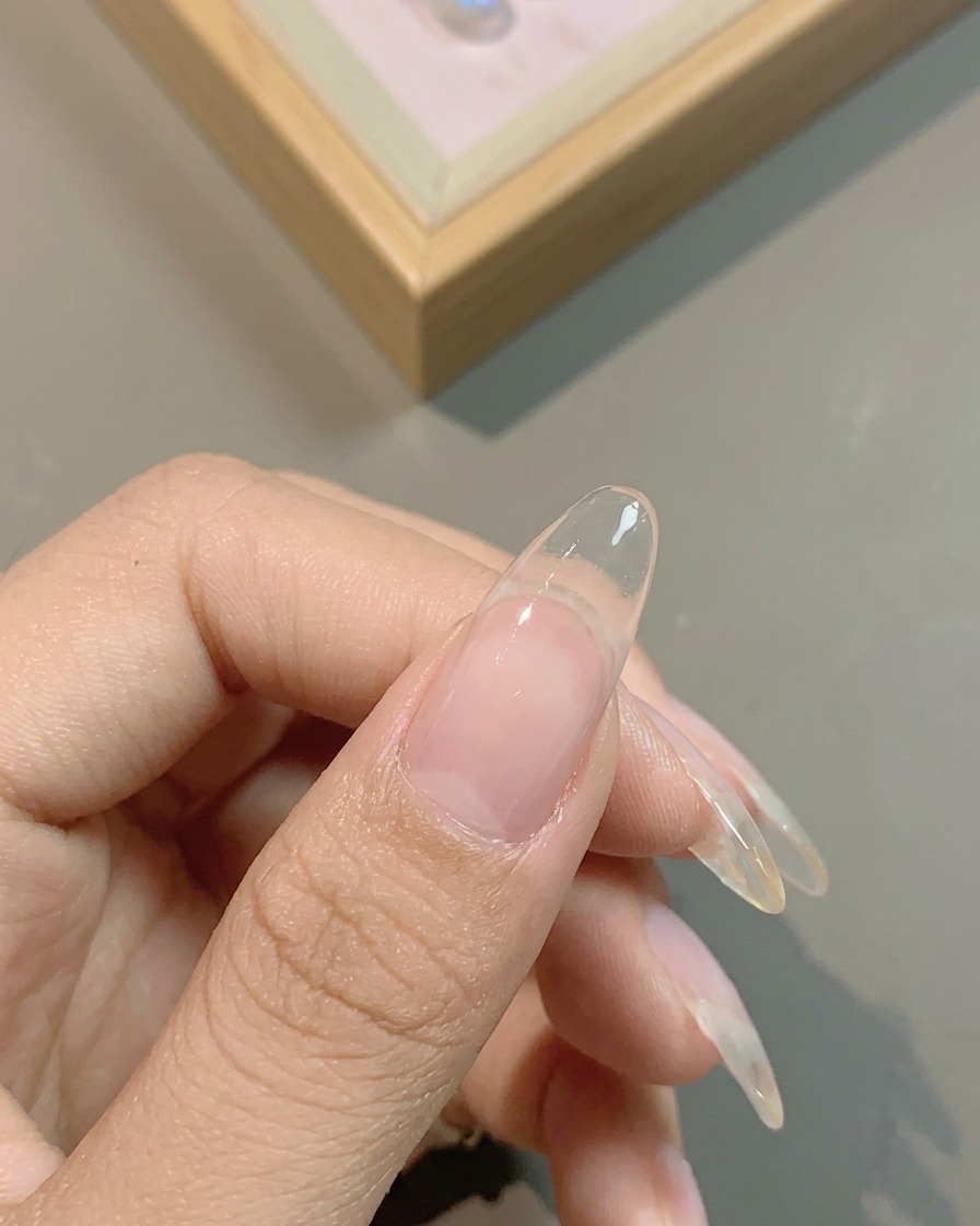 A brush applying a translucent milky white gel base coat over the entirety of a natural fingernail.