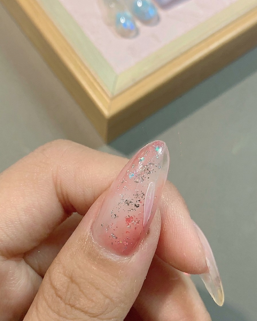 A close-up of a fine liner brush drawing delicate white flower petals over a pink, glitter-accented gel nail base.