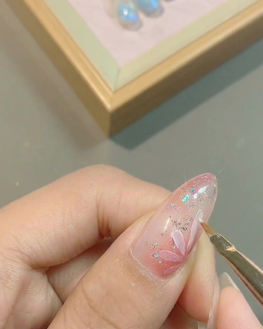 A close-up view of a brush precisely tracing the edges of white flower petals painted on a pink gel-coated fingernail.