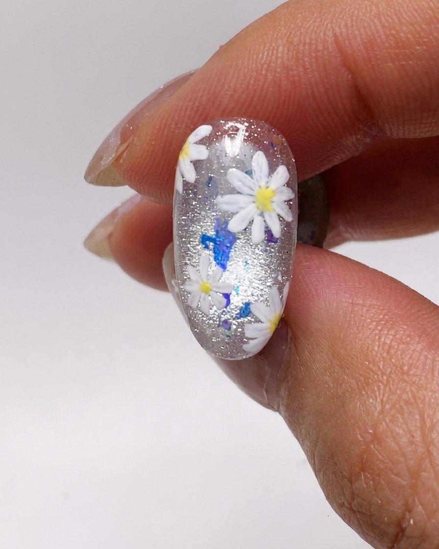 A close-up of a nail brush applying a thin coat of silver cat-eye gel polish to a natural fingernail.