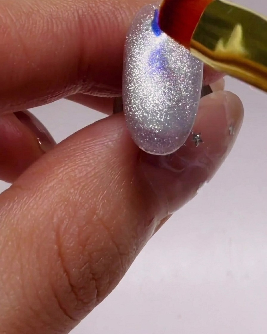 A small nail brush carefully placing iridescent blue and purple glitter flakes onto a shimmering silver nail.
